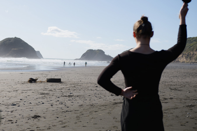 A person dressed in black, seen from behind, swings a poi on a beach. In the distance a person is lying on the sand, next to a large tractor tyre. 