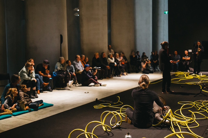 A musical performance of Weather Assemblage in the Len Lye Centre, featuring lots of curly yellow air hoses powering brass instruments. 