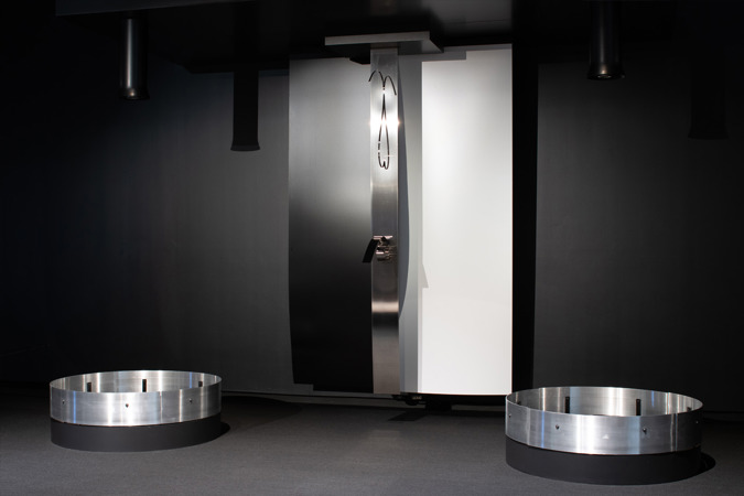 Len Lye's artwork storm, comprising three steel artworks - two sheets hanging from above, a strip of stainless steel in the centre, and two circular forms on the ground - exhibited in a black room. 